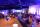 A party is taking place in a large room. Guests are gathered around tables holding glasses and chatting. Purple and blue light is cast throughout the room.
